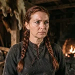 Femme viking forte et déterminée en tenue médiévale dans une longhouse scandinave, évoquant les épouses de chefs dans la série Vikings